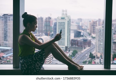 Woman Staying Home Using Mobile Phone By Window Indoors In The Dark. Asian Young Lady Pensive Looking At Cellphone Social Media App. Mental Health Online Addiction During Covid Confinement Concept.