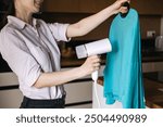 Woman steaming a blue shirt with handheld steamer at home. Garment care, laundry, wrinkle removal, home chores, clothing maintenance.