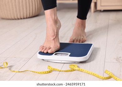 Woman stepping on floor scale and measuring tape at home, closeup. Weight control