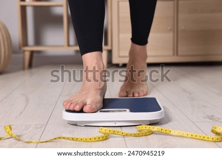 Woman stepping on floor scale and measuring tape at home, closeup. Weight control