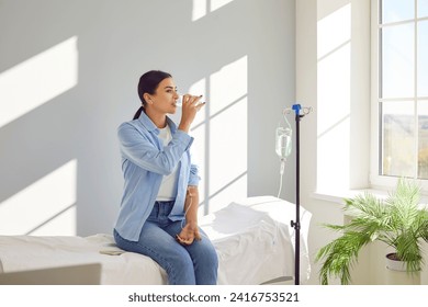 Woman with sterile tube in arm sitting by pole with IV bottle at medical center, drinking water and receiving vitamin therapy infusion to boost body immune system, preserve beauty or fight hangover - Powered by Shutterstock