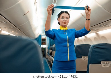 Mujeres azafatas con uniforme de azafata de aire sujetando el cinturón de seguridad mientras se paran cerca de los asientos de pasajeros en el salón de avión