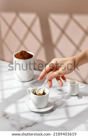Woman stir coffee with spoon. Coffee cup, hot drink in a mug. Beige and marble background with window shadow. Sunny, sunshine. 