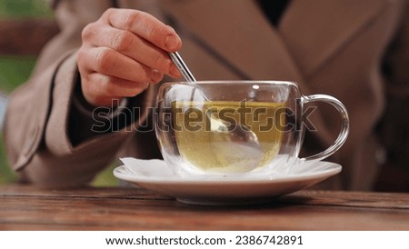 Woman stirs green tea with spoon in glass cup in cafe of park. Woman takes tea break with rotating spoon to stir sugar in cafe. Tea for maintaining woman figure with stirring spoon in cafe