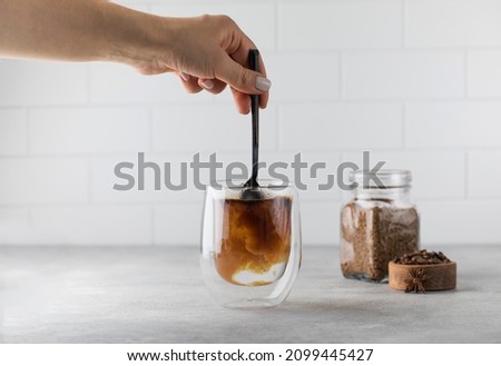 Woman stirs instant coffee in glass mug with boiled water on grey stone table