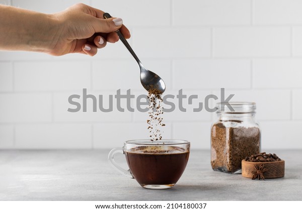 Woman stirs instant coffee in glass mug with boiled\
water on grey stone table