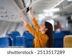 Woman Storing Luggage in Airplane Overhead Compartment: A young woman in a stylish mustard yellow jacket smiles as she stows her luggage in the overhead compartment of an airplane, a moment of travel 