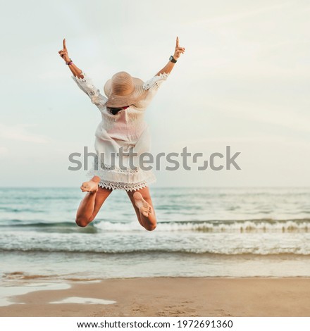 a woman with a straw hat jumps looking towards the sea on the beach shore. she is on her back. concept happiness and vacation.