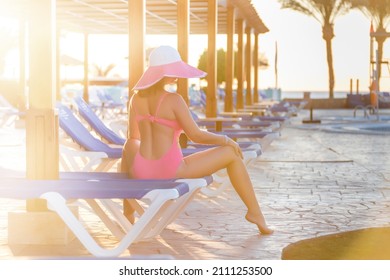 A Woman In A Straw Hat Is Relaxing On A Daybed Next To A Luxury Summer Pool In Egypt, Caribbean, Maldives, Italy, Spain, Florida, Manocco, Travel Concept Time. Palm Trees, Sea And Sun All Around.