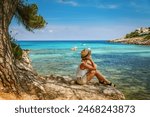 A woman in a straw hat sitting by the shoreline, admiring the clear blue sea, a perfect scene of Mallorca tourism