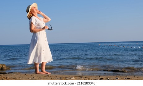 Woman In A Straw Hat And White Dress Takes Off Her Sunglasses And Enjoys The Sun While Walking Along The Seashore And Admires The Blue Sea On A Sunny Summer Day, Enjoying Freedom And Relaxation. The