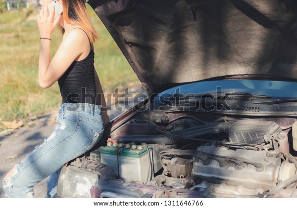  woman in\
street looking at broken down car engine on street. Opening hood\
and calling for help on cell\
phone