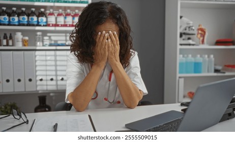 Woman stressed in clinic workplace with hands covering face by laptop and medical supplies in background. - Powered by Shutterstock