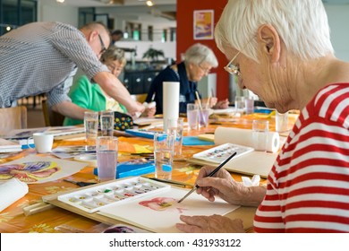 Woman in striped red and white shirt working on watercolor painting at table with other students in spacious studio - Powered by Shutterstock