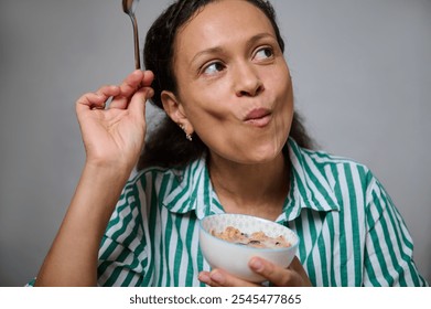 A woman in a striped shirt enjoys a healthy breakfast with a thoughtful expression, holding a bowl and spoon. - Powered by Shutterstock