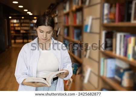 Woman student with a book is standing in a library or bookstore.  Girl reading a book in the library. Teaching students. Concept: education, university, student learning. Casual clothes, long hair