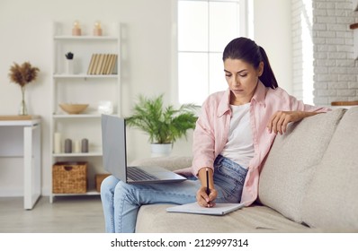 Woman Studying Online. Serious Focused Young Lady Sitting On Comfortable Sofa At Home With Laptop Computer On Her Lap, Using Modern Digital Learning Tools, Attending Webinar And Taking Notes On Paper