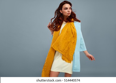 Woman Style And Fashion. Portrait Of Beautiful Sexy Model Girl In Stylish Fashionable Clothes, White Dress, Light Blue Coat Jacket, Yellow Scarf  Posing On Grey Background In Studio. High Resolution