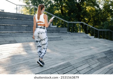 A woman in styling athletic wear leggings and bra walking on wooden pier outdoors. Fitness girl holding phone on her hands - Powered by Shutterstock