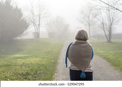 Woman in a stylish knitted cape and scarf combination walking along a misty winter road away from the camera in an healthy outdoor active lifestyle concept