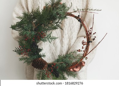 Woman in stylish knitted sweater holding modern christmas wreath on white background. Rustic christmas wreath in female hands with fir branches, berries and cone. Merry Christmas - Shutterstock ID 1816829276