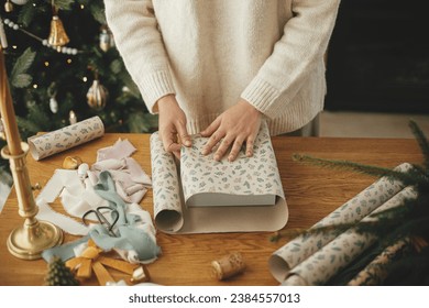 Woman in stylish sweater wrapping christmas gift in paper on wooden table with festive decorations in decorated scandinavian room. Merry Christmas! Hands packing present close up - Powered by Shutterstock