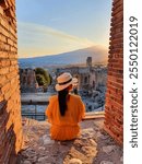 A woman in a stylish yellow dress and wide-brimmed hat sits quietly among ancient ruins, The sky glows softly as the silhouette of Mount Etna, Taormina, the ancient Greek theater in Sicily