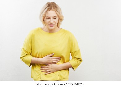 Woman With Suffer Expression On Face Holding Stomach And Squirming With Pain. Indigestion, Intestinal Problems, Gas And Bloating On A White Background