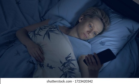 woman suffering from insomnia uses a smartphone while lying in bed in the dark.