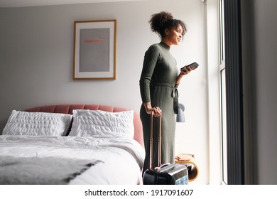 Woman With Suitcase Checking Out Of Boutique Hotel Standing By Bed Waiting For Taxi Ordered On Mobile Phone App