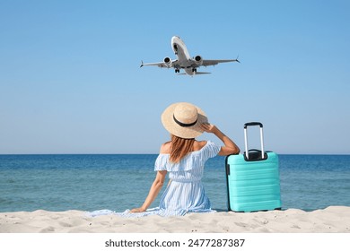 Woman with suitcase on sandy beach looking at airplane flying in sky, back view - Powered by Shutterstock