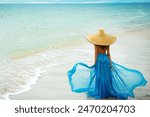 Woman in Summer Hat walking away on Beach. Beautiful Girl in Blue fluttering Dress looking at Sea. Holiday Travel. Tropic Vacations. Mind Relaxation and Meditation