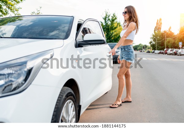 woman in summer opens car door,\
stands by car, a white sedan, a beautiful girl smiles, long hair\
sunglasses. Denim shorts and a white top. Parking by the\
road