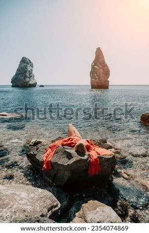 Woman summer travel sea. Happy tourist in long red dress enjoy taking picture outdoors for memories. Woman traveler posing on beach at sea surrounded by volcanic mountains, sharing travel adventure