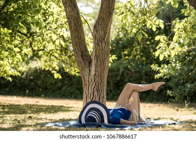 Woman In A Sun Hat Relaxing On A Grass In Summer Lying On A Rug In The Shade Of A Tree With A Refreshing Drink In Her Hand.