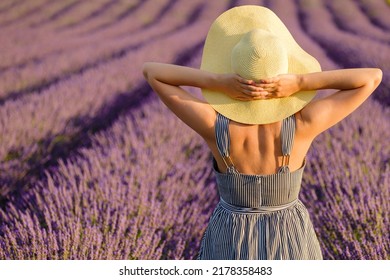 Woman In Sundress Puts Hands On Straw Hat Standing On Lavender Field. Young Lady Enjoys Beautiful Nature On Sunny Summer Day Backside View