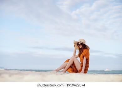 Woman In A Sundress Sits On The Sand On The Beach Hat On Her Head Side View                              