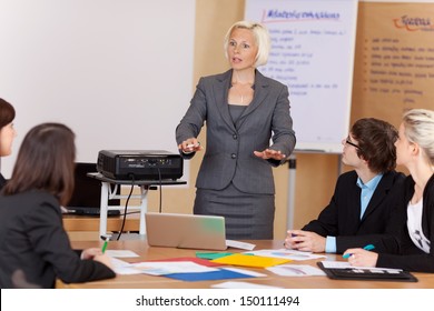 Woman Sung A Projector Giving A Corporate Training Class To A Group Of Young Businesspeople Around A Table Gesturing With Her Hands As She Explains Something