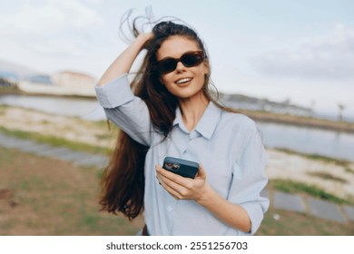 Woman, sunglasses, blue shirt, cell phone, water, outdoors a woman wearing sunglasses and a blue shirt is holding her cell phone near a body of water with another body of water and clear skies in the - Powered by Shutterstock