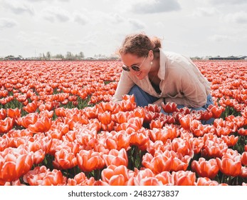 Woman in sunglasses and casual clothes, leaning forward to admire blooming red tulips in a vast field on a cloudy day - Concept of appreciating natural beauty and tranquility - Powered by Shutterstock