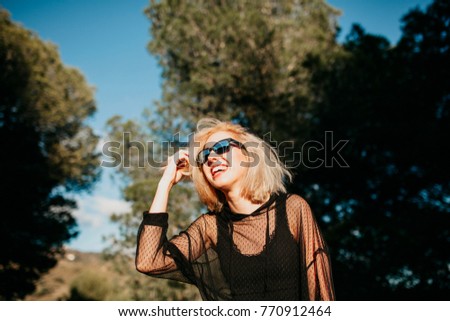 Kühle blonde Frau mit Sonnenbrille und schwarzem Kleid, die sich bei Sonnenuntergang in der Natur amüsiert.