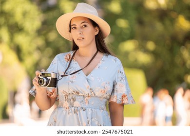 A woman in a sunhat and floral dress holds a vintage camera while looking directly at the camera. She stands outdoors with a backdrop of blurred greenery, highlighting the serene environment.  - Powered by Shutterstock