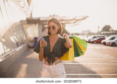 Woman At Sunset With Colorful Shopping Bags And Parking Lot By Shopping Mall Happy With Mobile Phone