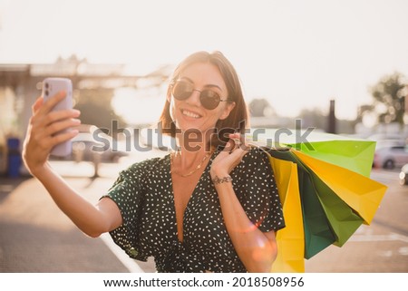 Woman at sunset with colorful shopping bags and parking lot by shopping mall happy with mobile phone take photo selfie