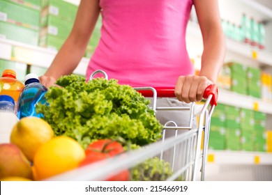 Woman at supermarket pushing a shopping cart filled with fresh fruit and vegetables.