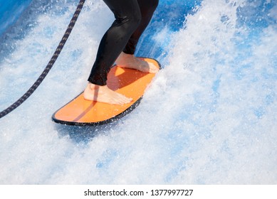 Woman Surfing In Beach Wave Simulator Attraction Of Water Park, Wearing Black Swiming Suit Balancing On Orange Surfboard In Fake Wave.  Outdoor Water Sport Activity.