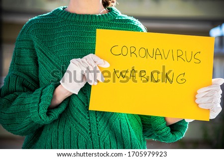 Woman with surgical latex gloves holding yellow paper sign with coronavirus warning. Covid-19 pandemic.