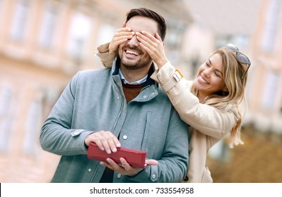 Woman Surprising His Boyfriend With A Gift.Young Woman With Giftbox Closing His Boyfriend Eyes To Make A Surprise For Him.