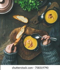 Woman In Sweater Eating Corn Creamy Soup With Shrimps In Individual Pot, Top View. Woman' S Hand Keeping Spoon And Bread Slice. Flat-lay Of Rustic Dinner Table. Slow Food, Winter Warming Food Concept