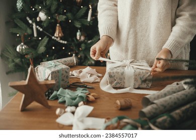 Woman in sweater wrapping christmas gift with stylish ribbon on wooden table with festive decorations in decorated scandinavian room. Merry Christmas! Hands packing present close up - Powered by Shutterstock